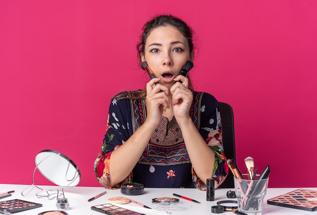 Surpris jeune fille brune assise à table avec des outils de maquillage tenant des pinceaux de maquillage