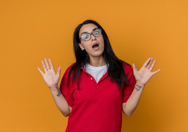 Surpris jeune fille caucasienne brune à lunettes optiques portant une chemise rouge lève les mains à la recherche d'isolement sur le mur orange