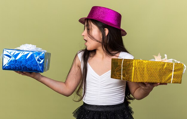 surpris jeune fille caucasienne avec un chapeau de fête violet regardant une boîte-cadeau tenant sur chaque main isolée sur un mur vert olive avec espace de copie