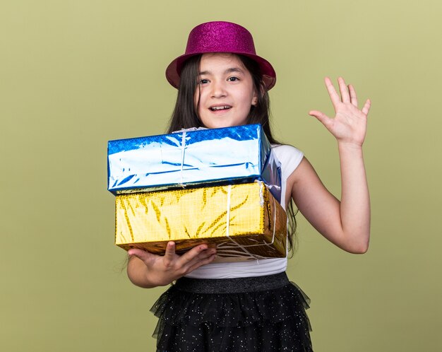 surpris jeune fille caucasienne avec chapeau de fête violet tenant des coffrets cadeaux et debout avec la main levée isolée sur un mur vert olive avec espace de copie