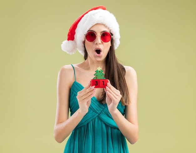 Surpris jeune fille caucasienne à lunettes de soleil avec bonnet de noel tenant ornement d'arbre de Noël