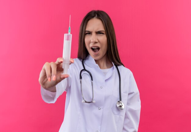 Surpris jeune fille médecin portant stéthoscope robe médicale regardant la seringue dans sa main sur fond rose isolé