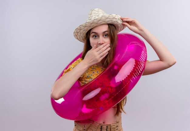 Surpris jeune fille portant chapeau et anneau de bain mettant les mains sur la bouche et le chapeau sur un mur blanc isolé avec copie espace