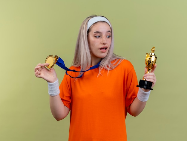 Surpris, une jeune fille sportive caucasienne avec des bretelles portant un bandeau et des bracelets détient une médaille d'or et regarde la coupe du vainqueur