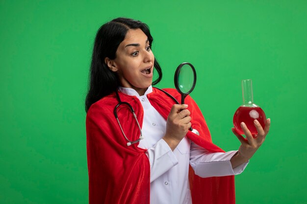 Surpris jeune fille de super-héros portant une robe médicale avec stéthoscope tenant et regardant la bouteille en verre de chimie remplie de liquide rouge avec loupe isolé sur vert