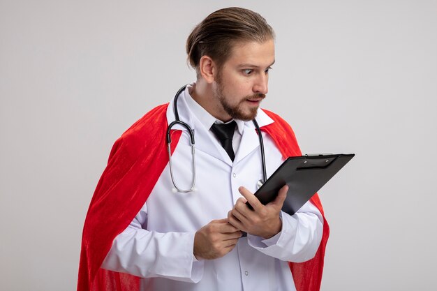 Surpris jeune gars de super-héros portant une robe médicale avec stéthoscope tenant et regardant le presse-papiers isolé sur fond blanc