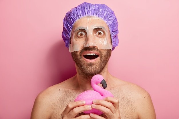 Surpris jeune homme applique un masque cosmétique détient une éponge de bain en forme de flamant rose, porte une bonnet de douche, a une routine d'hygiène, se tient avec le torse nu