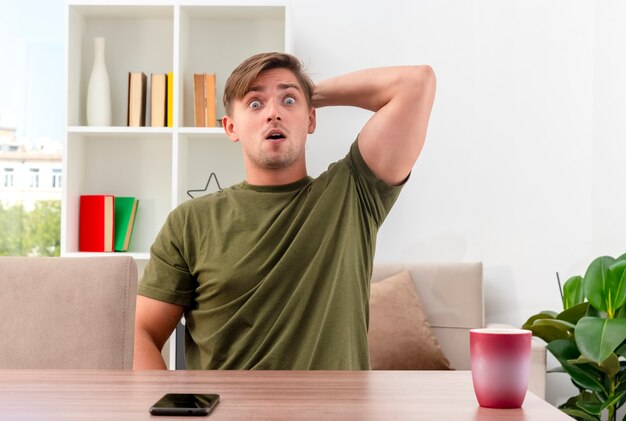 Surpris jeune homme beau blond est assis à table avec téléphone et tasse mettant la main sur la tête derrière en regardant la caméra à l'intérieur du salon