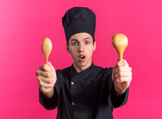 Surpris jeune homme blond cuisinier en uniforme de chef et casquette regardant la caméra étirant des cuillères en bois vers la caméra isolée sur un mur rose