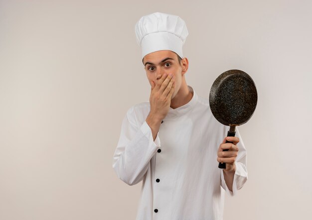 Surpris jeune homme cuisinier portant un uniforme de chef bouche couverte avec la main et tenant une poêle avec espace copie