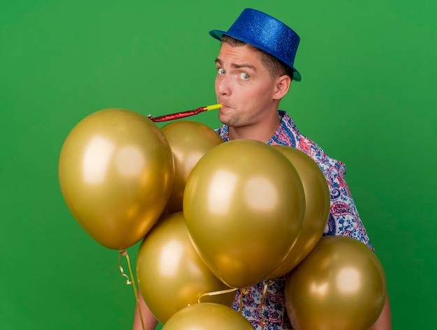 Photo gratuite surpris jeune homme de fête portant un chapeau bleu soufflant du parti debout derrière des ballons isolés sur le vert