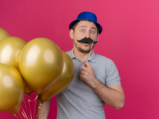 Surpris jeune homme de fête portant un chapeau bleu tenant des ballons avec une fausse moustache sur bâton isolé sur rose