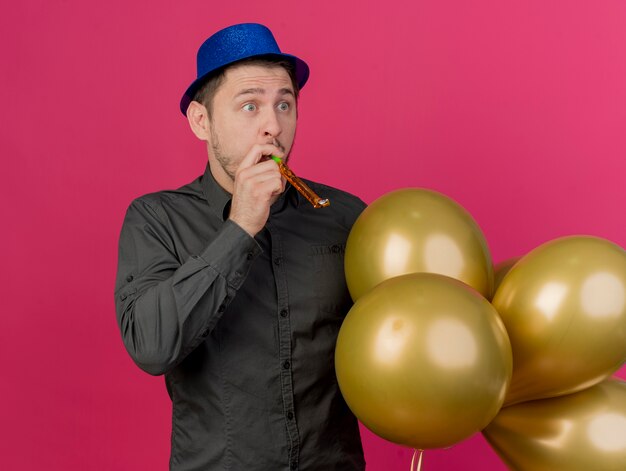 Surpris jeune homme de fête portant un chapeau bleu tenant des ballons soufflant sifflet isolé sur rose