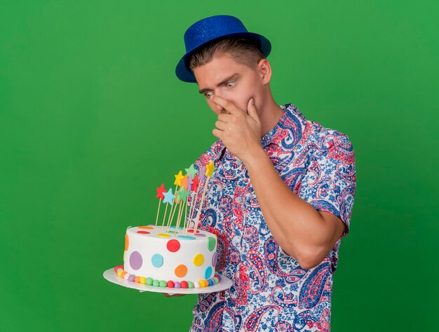 Surpris jeune homme de fête portant un chapeau bleu tenant et regardant le gâteau mettre la main sur la bouche isolé sur vert