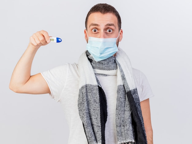 Surpris jeune homme malade portant un masque médical et une écharpe tenant un thermomètre isolé sur fond blanc