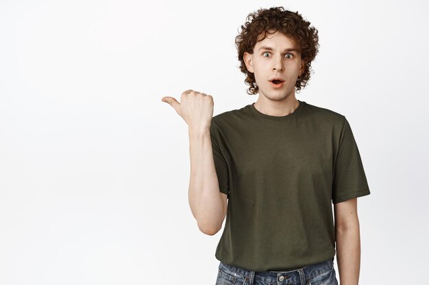 Surpris jeune homme pointant vers la gauche regardant impressionné par la caméra vérifiant la publicité montrant l'offre promotionnelle de l'offre de magasin debout sur fond blanc