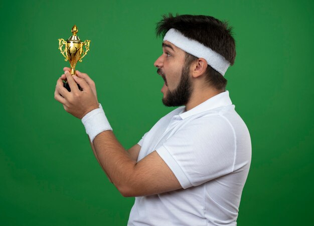 Surpris jeune homme sportif debout en vue de profil portant bandeau et bracelet tenant et regardant la coupe du gagnant isolé sur le mur vert