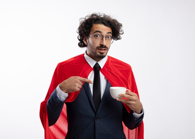 Surpris jeune homme super-héros caucasien dans des lunettes optiques portant un costume avec une cape rouge tient et pointe vers la tasse