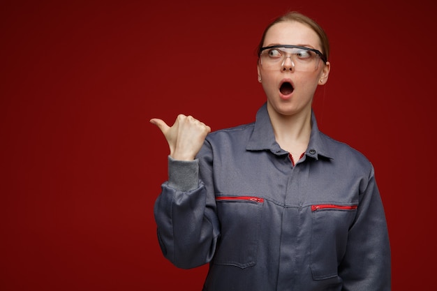 Surpris jeune ingénieur blonde portant des lunettes de sécurité et uniformes à la recherche et pointant sur le côté