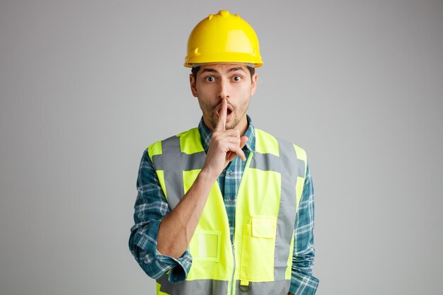 Surpris jeune ingénieur masculin portant un casque de sécurité et un uniforme regardant la caméra montrant un geste silencieux isolé sur fond blanc