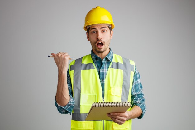 Surpris jeune ingénieur masculin portant un casque de sécurité et un uniforme tenant un bloc-notes et un crayon regardant la caméra pointant vers le côté isolé sur fond blanc