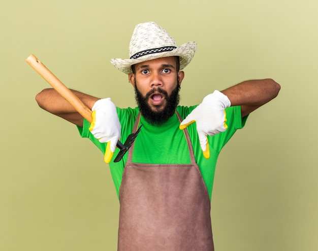 Surpris jeune jardinier afro-américain portant un chapeau de jardinage avec des gants tenant des pointes de râteau vers le bas isolé sur un mur vert olive