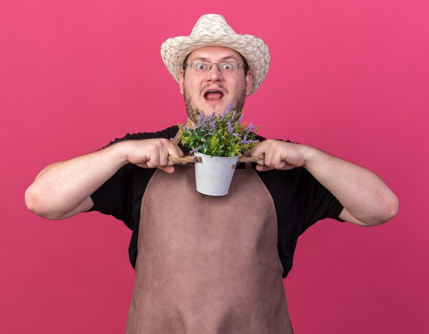 Surpris jeune jardinier mâle portant chapeau de jardinage tenant une fleur en pot de fleurs isolé sur mur rose