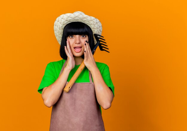 Surpris, une jeune jardinière en uniforme portant un chapeau de jardinage met les mains sur le visage