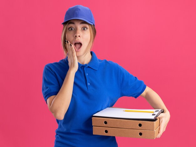 Surpris jeune jolie femme de livraison en uniforme met la main sur le visage et détient le presse-papiers de boîtes à pizza isolé sur mur rose