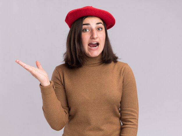 Surpris jeune jolie fille caucasienne avec chapeau de béret pointant sur le côté avec la main isolée sur mur blanc avec espace de copie