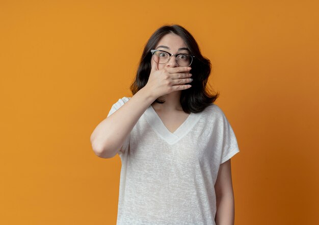 Surpris jeune jolie fille caucasienne portant des lunettes mettant la main sur la bouche isolé sur fond orange avec copie espace