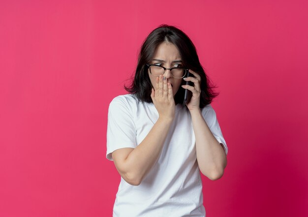 Surpris jeune jolie fille caucasienne portant des lunettes regardant côté parler au téléphone mettant la main sur la bouche isolé sur fond cramoisi avec espace copie