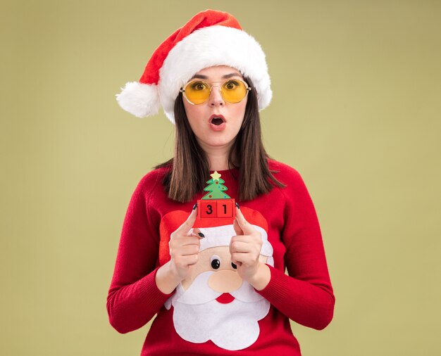 Surpris jeune jolie fille caucasienne portant pull du père noël et bandeau avec des lunettes tenant jouet arbre de Noël avec date regardant la caméra isolée sur fond vert olive