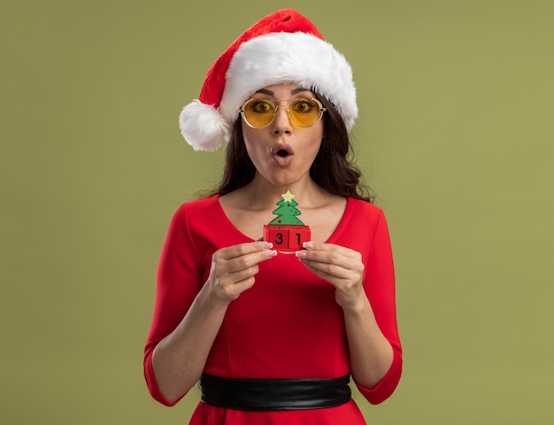 Surpris jeune jolie fille portant un bonnet de noel et des lunettes tenant un jouet d'arbre de noël avec une date isolée sur un mur vert olive