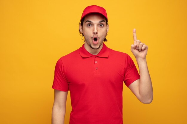 surpris jeune livreur portant l'uniforme et la casquette regardant la caméra pointant le doigt vers le haut isolé sur fond jaune