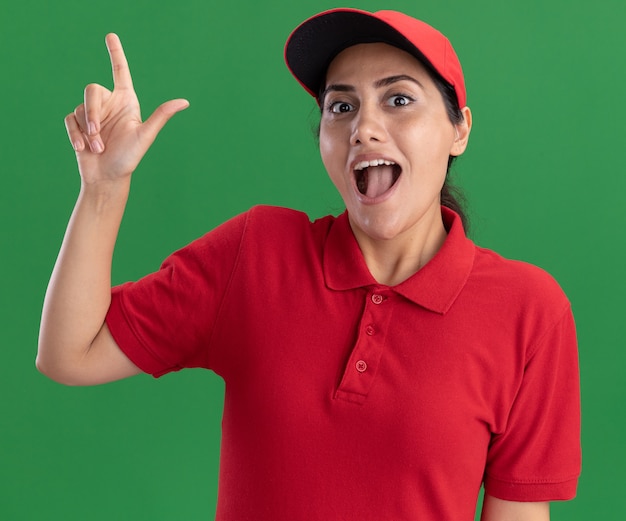 Surpris jeune livreuse portant l'uniforme et les points de casquette à up isolé sur mur vert