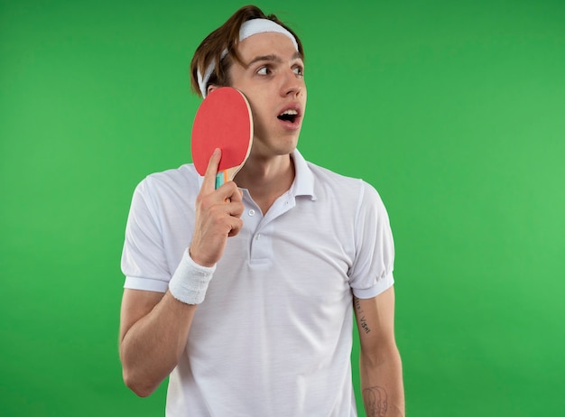 Photo gratuite surpris jeune mec sportif regardant côté portant un bandeau avec bracelet mettant la raquette de ping-pong au visage isolé sur mur vert