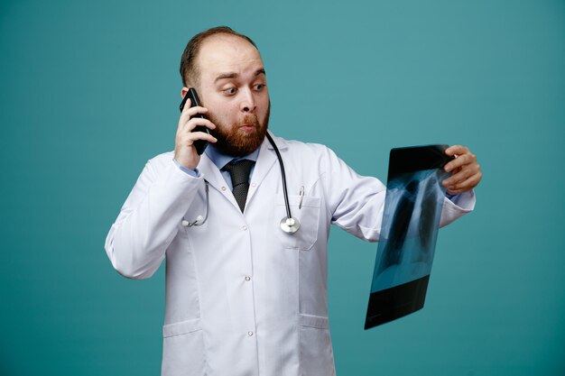 Surpris jeune médecin de sexe masculin portant un manteau médical et un stéthoscope autour de son cou tenant et regardant une radiographie tout en parlant au téléphone isolé sur fond bleu