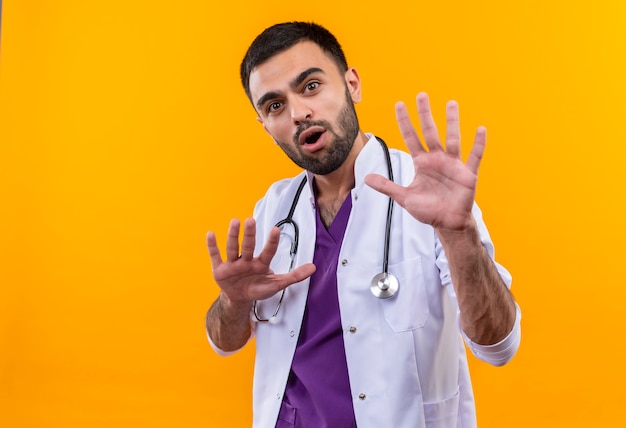 Surpris jeune médecin de sexe masculin portant une robe médicale stéthoscope main levée sur mur jaune isolé