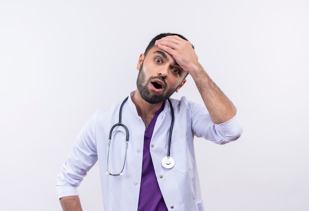 Surpris jeune médecin de sexe masculin portant une robe médicale stéthoscope a mis sa main sur le front sur blanc isolé