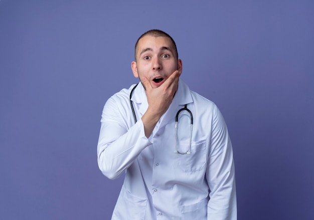 Surpris jeune médecin de sexe masculin portant une robe médicale et un stéthoscope tenant son menton isolé sur un mur violet