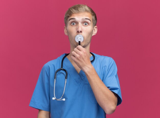 Surpris jeune médecin de sexe masculin portant l'uniforme de médecin avec stéthoscope mettant le stéthoscope sur la bouche isolé sur le mur rose