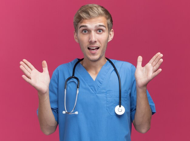 Surpris jeune médecin de sexe masculin portant l'uniforme de médecin avec stéthoscope répandant les mains isolé sur mur rose