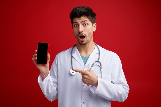 Surpris jeune médecin de sexe masculin portant un uniforme médical et un stéthoscope autour du cou regardant la caméra montrant un téléphone portable pointant dessus isolé sur fond rouge