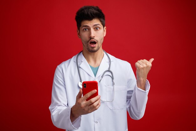 Surpris jeune médecin de sexe masculin portant un uniforme médical et un stéthoscope autour du cou tenant un téléphone portable regardant la caméra pointant vers le côté isolé sur fond rouge