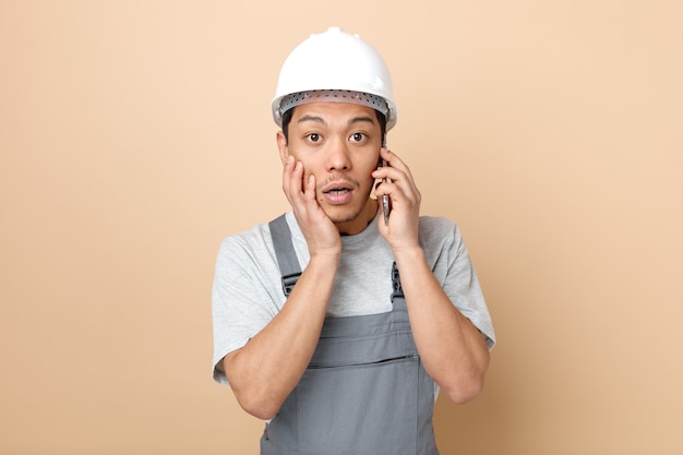 Surpris jeune travailleur de la construction portant un casque de sécurité et un uniforme parlant au téléphone en gardant la main sur le visage