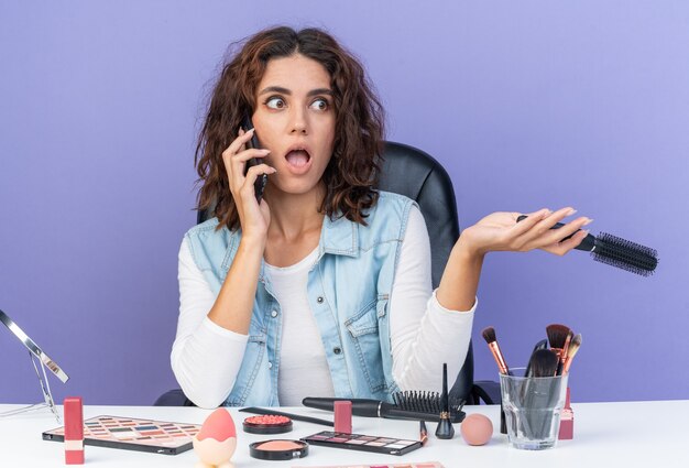 Surpris jolie femme caucasienne assise à table avec des outils de maquillage parlant au téléphone et tenant un peigne regardant de côté