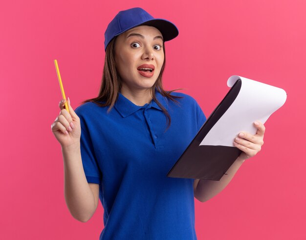 Surpris jolie femme de livraison en uniforme tenant un crayon et un presse-papiers sur rose