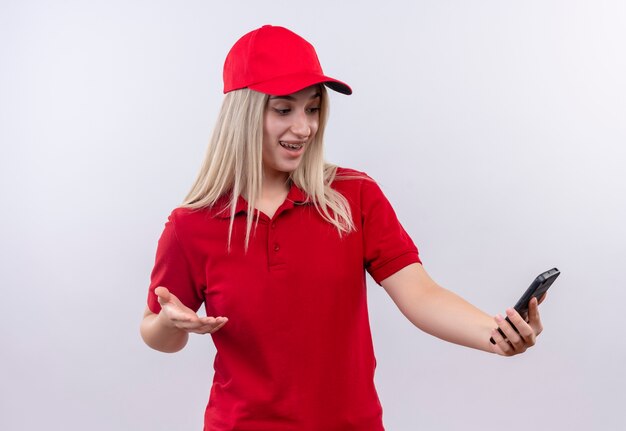 Surpris livraison jeune fille portant un t-shirt rouge et une casquette regardant le téléphone sur sa main sur fond blanc isolé