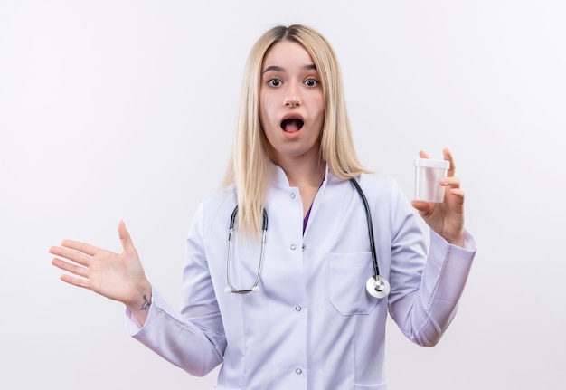 Surpris médecin jeune fille blonde portant un stéthoscope et une robe médicale tenant vide peut levé la main sur fond blanc isolé